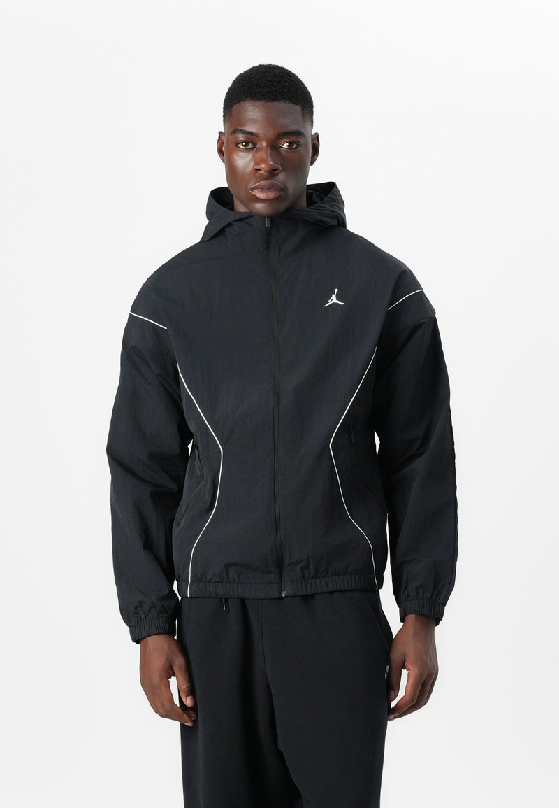 Veste coupe-vent noire avec capuche, dotée d'accents blancs et d'un logo Jordan sur la poitrine. Poignets et taille élastiques. Matériau léger.