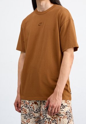 Personne portant un t-shirt Nike marron et un short beige avec des motifs abstraits marron, noir et orange, debout devant un fond uni.