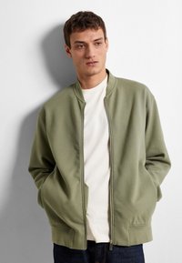 Veste bomber verte clair avec fermeture éclair à l'avant, col montant et poches latérales. Portée sur un T-shirt crème, associée à un jean foncé.