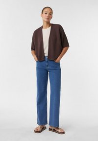 Bruine gebreide cardigan met open voorkant, gedragen over een crèmekleurig topje. Combinatie met blauwe rechtne pijpen jeans en zwarte sandalen met zilveren accenten.