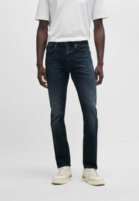 Dark blue denim jeans with a slim fit, featuring a classic five-pocket design and a subtle fade. Worn with a plain white t-shirt and beige sneakers.