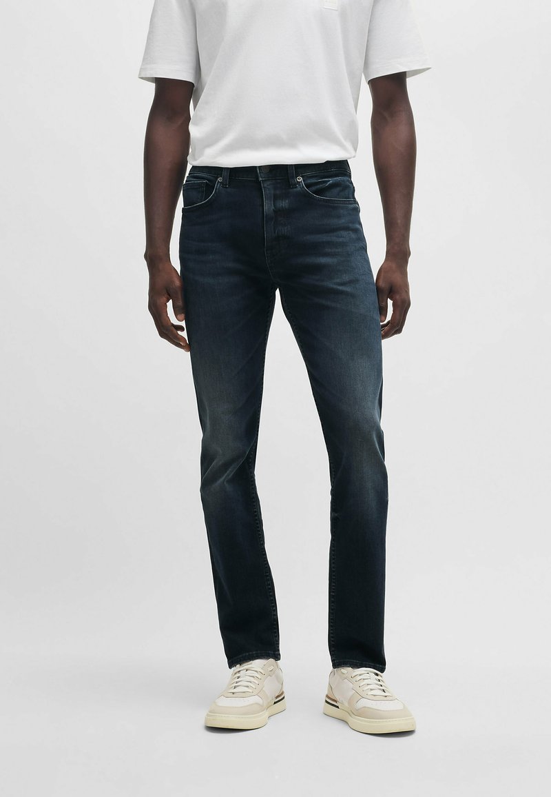 Dark blue denim jeans with a slim fit, featuring a classic five-pocket design and a subtle fade. Worn with a plain white t-shirt and beige sneakers.