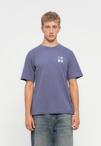Joven con cabello rubio corto, vistiendo una camiseta púrpura holgada con caracteres asiáticos blancos y rojos en el pecho izquierdo y jeans azules desgastados.