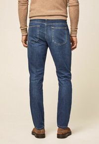 Hombre vestido con jeans azul ajustados, camisa de manga larga beige y zapatos de ante marrones, de pie contra un fondo neutro.