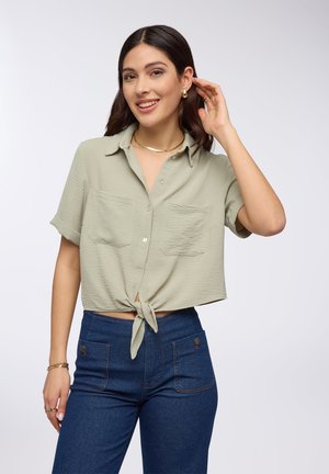 Camicia con nodo anteriore in tessuto verde chiaro con finitura testurizzata, due tasche sul petto, maniche corte, abbinata a jeans blu a vita alta.