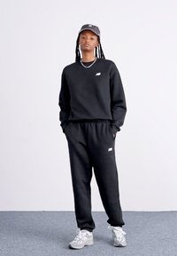 Sort sweatshirt og sweatpants med elastisk talje, der har et hvidt logo på brystet. Grå sneakers med snørebånd fuldender outfittet.