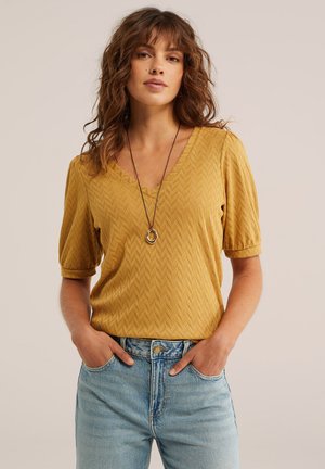 Frau mit welligem braunem Haar, die ein senfgelbes, strukturiertes V-Ausschnitt-Oberteil, blaue Jeans und eine lange Anhänger-Halskette trägt, Hände in den Taschen.