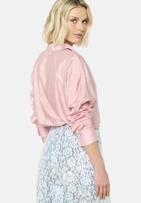 Lichtroze satijnen blouse met een kraag, verlaagde schouders en gathered manchetten, gecombineerd met witte bloemenkant broeken. Zichtbaar achterkant detail.