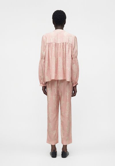 Άτομο που στέκεται με την πλάτη γυρισμένη, φοράει φαρδιά ροζ μπλούζα με μοτίβο παϊνσλεϊ και αντίστοιχο φαρδύ παντελόνι με μαύρα παπούτσια σε λευκό φόντο.