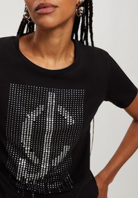 Czarna koszulka z nadrukiem z przodu, w którym srebrne cyrkonie ułożone są w kształt centralnej grafiki. Okrągły dekolt i krótkie rękawy.