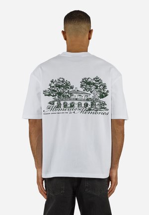 Άνδρας που φοράει λευκό μπλουζάκι με μαύρη μελάνη απεικόνιση ενός σπιτιού και δέντρων, με την επιγραφή "Στιγμές για Αναμνήσεις" στην πλάτη.