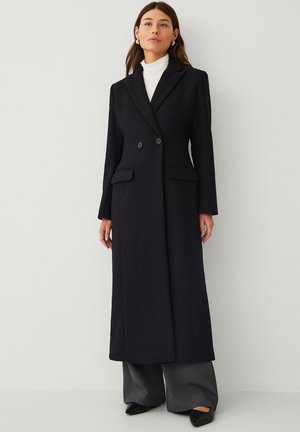 Woman wearing a long black double-breasted coat over a white turtleneck, gray wide-leg pants, and black pointed shoes standing against a plain wall.