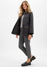 Zwarte gewatteerde jas met gouden accenten, voorzien van een kraag en zakken. Draag het over een grijze trui en een genoteerde broek met zwarte instappers.