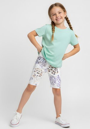 Uśmiechnięta dziewczyna z warkoczami, ubrana w miętową koszulkę, białe szorty w kwiatowy wzór oraz białe trampki, stojąca z rękami na biodrach.