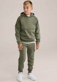 Olíva zöld kapucnis pulóver bordázott részletekkel és logóval, hozzáillő zsebes melegítőnadrággal, bokáig érő szegéllyel, fehér sportcipővel viselve.