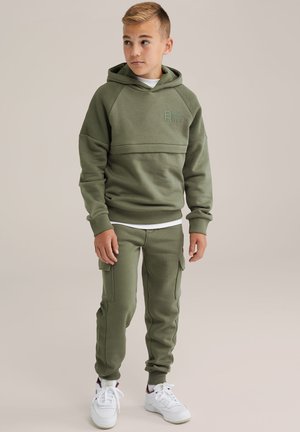 Felpa con cappuccio verde oliva con dettagli a coste e logo, abbinata a pantaloni jogger coordinati con tasche e caviglie a costine, indossati con sneaker bianche.