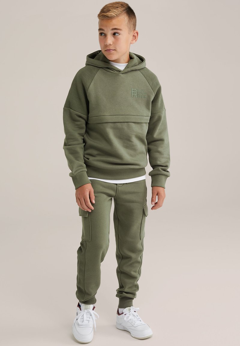Olíva zöld kapucnis pulóver bordázott részletekkel és logóval, hozzáillő zsebes melegítőnadrággal, bokáig érő szegéllyel, fehér sportcipővel viselve.