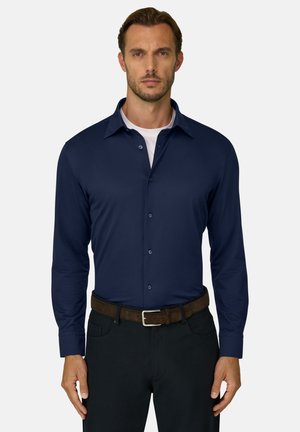 Homme aux cheveux courts portant une chemise bleu marine boutonnée sur un sous-vêtement blanc, un pantalon noir et une ceinture marron, debout devant un fond uni.