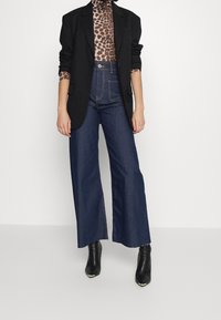 Pantalones de mezclilla oscuros de pierna ancha combinados con una blazer negra y un cuello de tortuga de estampado de leopardo. El material parece suave; los pantalones tienen una cintura alta.