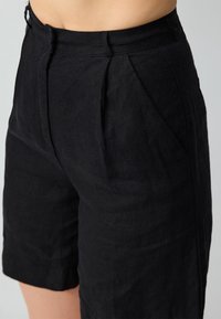 Pantaloni corti in lino nero con una vestibilità comoda, dotati di pieghe frontali, tasche laterali e una texture liscia. Rifiniti appena sopra il ginocchio.