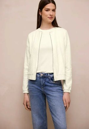 Vrouw draagt een witte, gestructureerde jas met rits over een wit topje en blauwe, hooggetailleerde jeans, staand tegen een beige achtergrond.