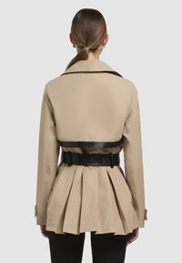 Beige trenchcoat met riem en geplooide achterkant, zwarte leren accenten en een kraag. Bevat een logo op de riem. Op maat gemaakt, middenlange pasvorm.