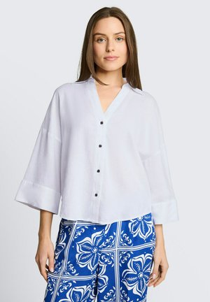Kobieta w luźnej białej bluzce zapinanej na guziki z szerokimi rękawami oraz niebieskich spodniach we wzory z dużymi motywami kwiatowymi.