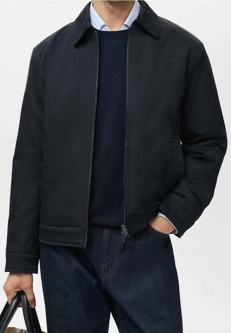 Homme portant une veste noire zippée sur un pull bleu marine et une chemise bleue, tenant un sac noir dans la main gauche, main droite dans la poche du jean.