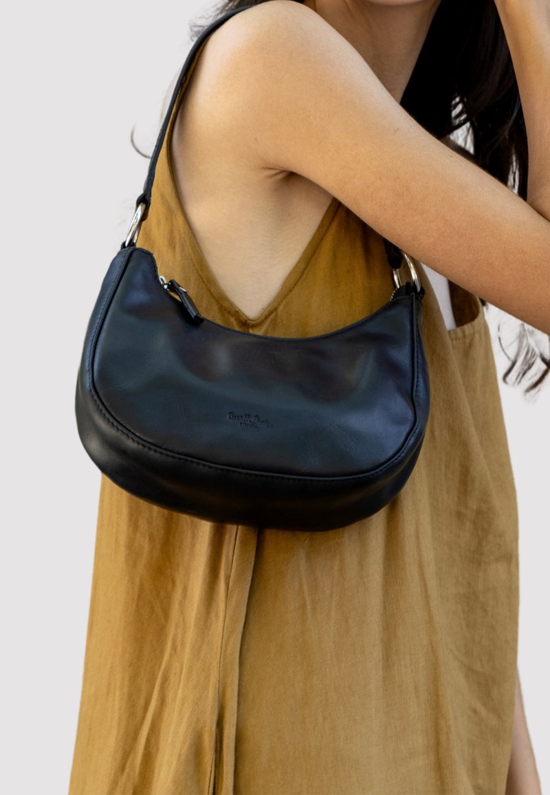 Juodas odinis peties krepšys su kreiva forma, turintis sidabrinę sagtį ir lygų paviršių, dėvimas ant rudos aprangos.