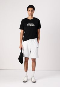 Svart t-skjorte med "Jack & Jones"-logo, lysegrå shorts, svart veske og hvite sokker med sko som har brune detaljer. Enkelt design og avslappet passform.