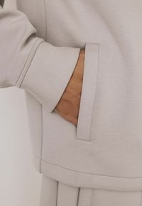 Light gray sweatshirt fabric with a smooth texture. Features a side pocket, ribbed cuffs, and visible stitching details.