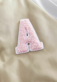 Beige tygbakgrund med ett texturerat rosa filtbrev "A" som kantas av vit tyg. Brevet har ett utskuret mönster i mitten.