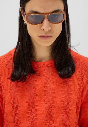 Persona con lunghi capelli scuri che indossa occhiali da sole con montatura color ambra, un piccolo orecchino e un maglione arancione brillante con tessitura, su uno sfondo chiaro uniforme.