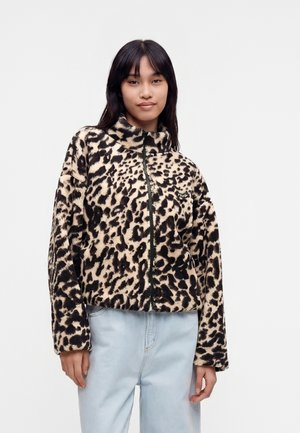 Giovane donna che indossa una giacca in pile beige e nera con stampa leopardata e jeans azzurri, in piedi davanti a uno sfondo semplice.