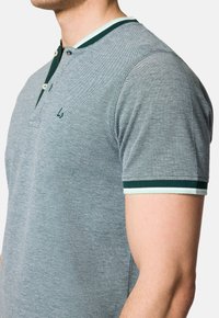 Szara koszulka polo z teksturowanym wykończeniem, zielonym i białym kołnierzykiem oraz akcentami na rękawach. Posiada dwa guziki z przodu i małe logo na piersi.