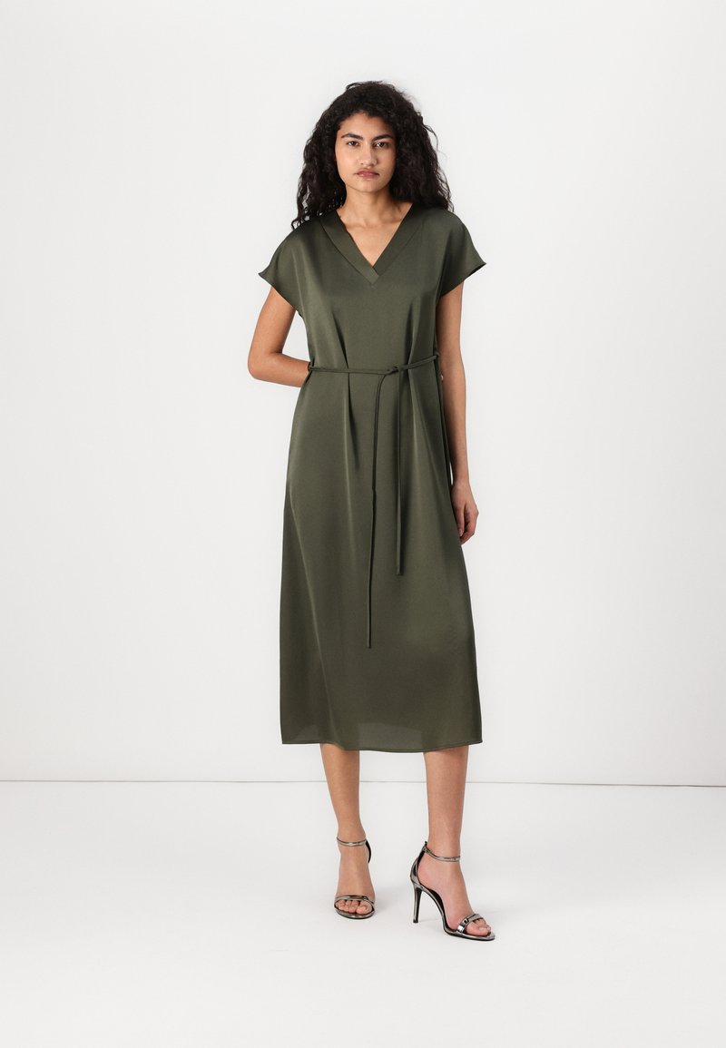 Vestido midi verde oliva con escote en V, mangas cortas y detalle de cintura atada. Tela suave con una silueta relajada. Tacones negros con tiras.