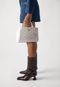 Bolso acolchado blanco con herrajes plateados, que presenta un candado y un logo. Se combina con botines de cuero marrón con tacones de bloque.