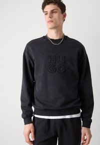 Czarna bluza z haftowanym logo "HUGO", ściągacze na rękawach i u dołu, luźny krój, noszona na białej warstwowej koszuli, teksturalny detal materiału.