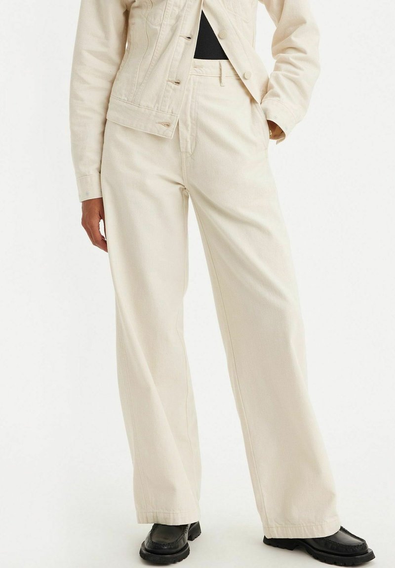 Femme portant un pantalon large couleur crème, une veste assortie, un haut noir et des chaussures noires, debout avec une main dans la poche.