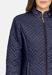 Chaqueta acolchada azul marino con cuello alto, cremallera frontal y un sutil patrón geométrico. Tejido suave con brillo delicado y detalles de broche.
