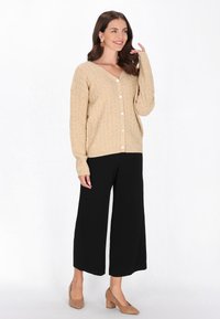 Beżowy sweter z dzianiny, z dekoltem w kształcie litery V i dużymi guzikami, zestawiony z czarnymi spodniami o szerokich nogawkach oraz beżowymi butami na słupku.
