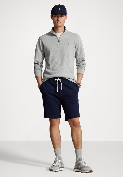 Grijze zip-nek pullover, marineblauwe shorts met drawstring, grijze sneakers en een marineblauwe pet met logo. Casual sportoutfit met gestructureerde materialen.