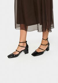 Zwarte lakleren schoenen met blokhak, een vierkante neus en meerdere enkelbandjes versierd met gouden hardware accenten.