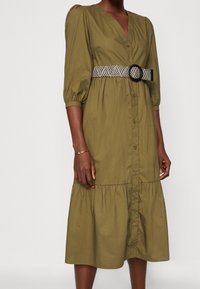 Oliwkowa sukienka zapinana na guziki z warstwowym dołem, bufkami i plecionym czarno-białym paskiem z okrągłą klamrą.