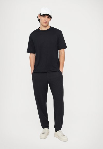 Czarny t-shirt z kr�ótkim rękawem oraz dopasowane dresy, oba wykonane z teksturowanego materiału. Model ma na sobie białe trampki i białą czapkę z logo.