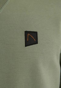 Pull vert clair avec un col en V, un tissu texturé, et un patch noir avec un motif géométrique orange.