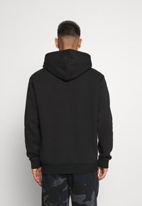 Schwarzer Hoodie mit weicher Textur, versehen mit einer großen Kapuze, Rippabschlüssen und einem geraden Saum, getragen über camouflagemusterte Hosen.