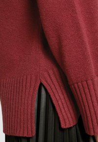 Pull en laine rouge tricoté avec des poignets côtelés et des fentes sur les côtés. Fabriqué en tissu doux, il présente une finition texturée et une coupe décontractée.