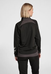 Sort atletisk jakke med høj krave, mesh-detaljer på skuldrene, ribbet kant og grå detaljer på ærmerne, designet for optimal komfort.