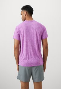 Lila gemustertes Kurzarm-Sportshirt mit rundem Hals und seitlichen Schlitzen, kombiniert mit grauen Shorts, von hinten betrachtet.